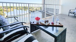 A balcony with a view of a body of water, a white railing, a black and white striped sofa, a small table with a vase of flowers, a pair of sunglasses, and two bottles of water. - Light Fountain Show View, Laketown  735, Spectacular Unit And Amenities (Panama City Beach)