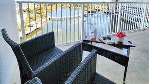 Un balcon agrémenté de meubles en osier, une table avec un vase de fleurs et une vue sur une étendue d’eau et des bâtiments.