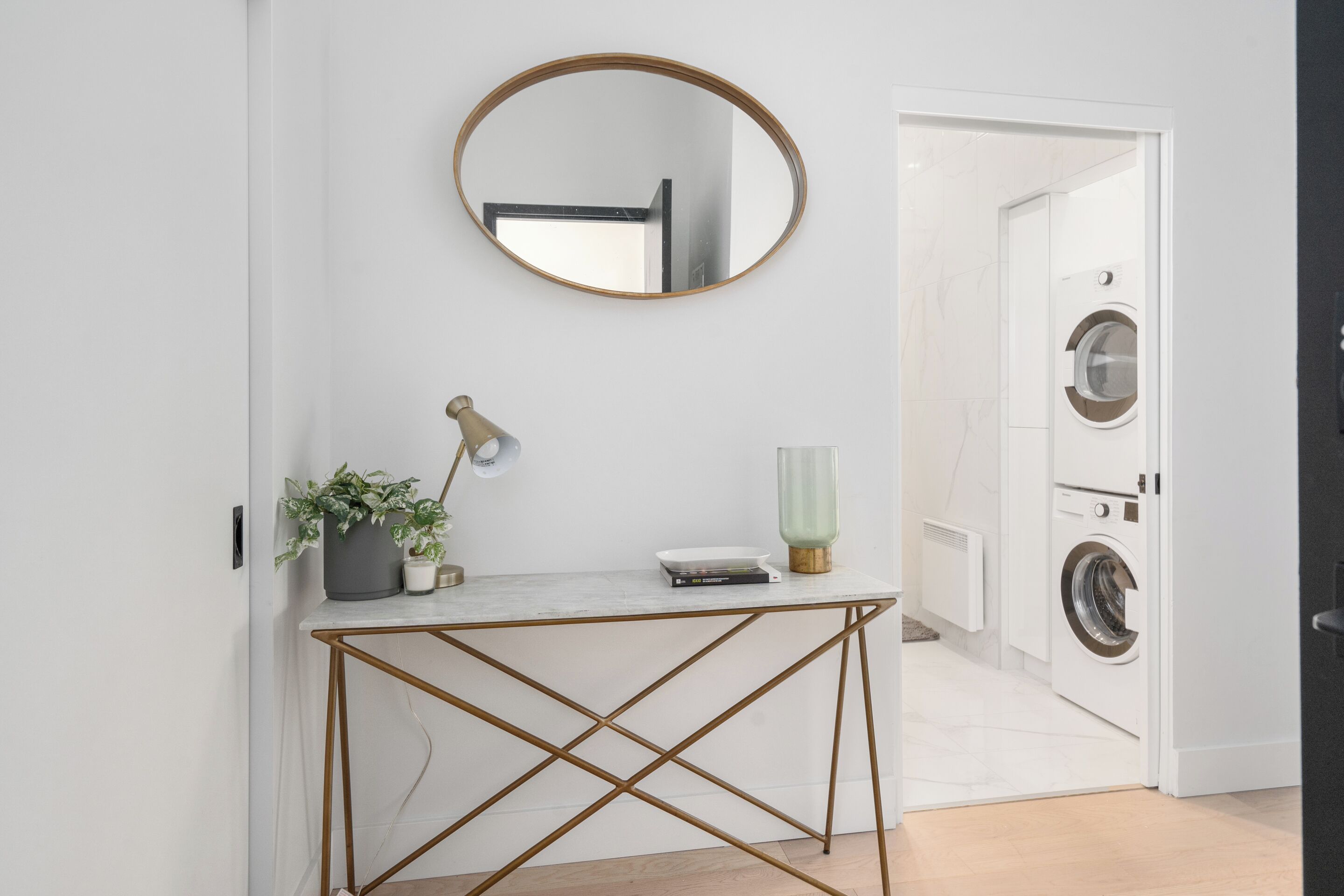 Een moderne badkamer met een wit wastafelmeubel, een ronde spiegel, een plant in een pot, een lamp en een wasmachine op de achtergrond.