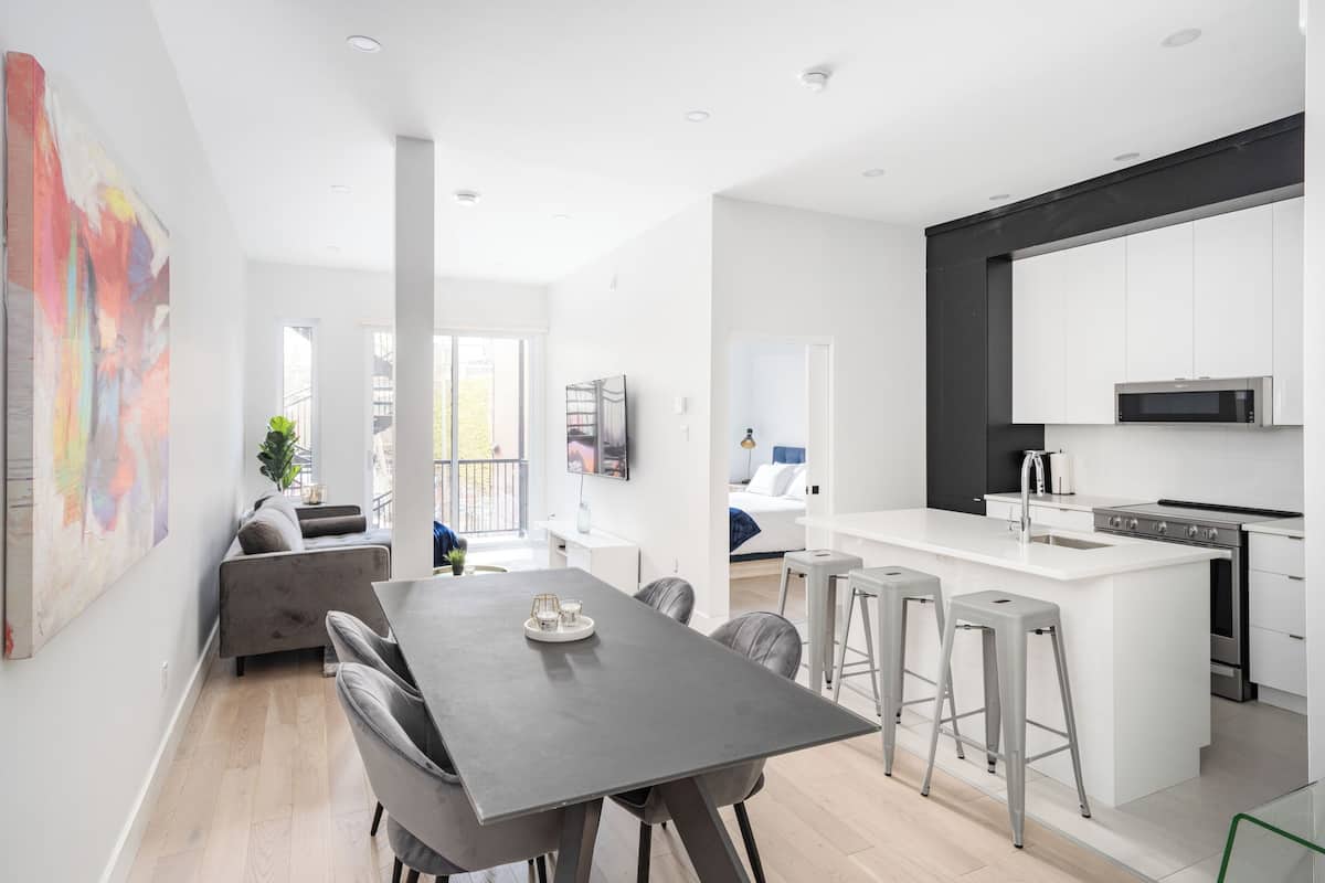 A modern kitchen with white cabinets, a large island with stools, and a dining area with a grey table and chairs.