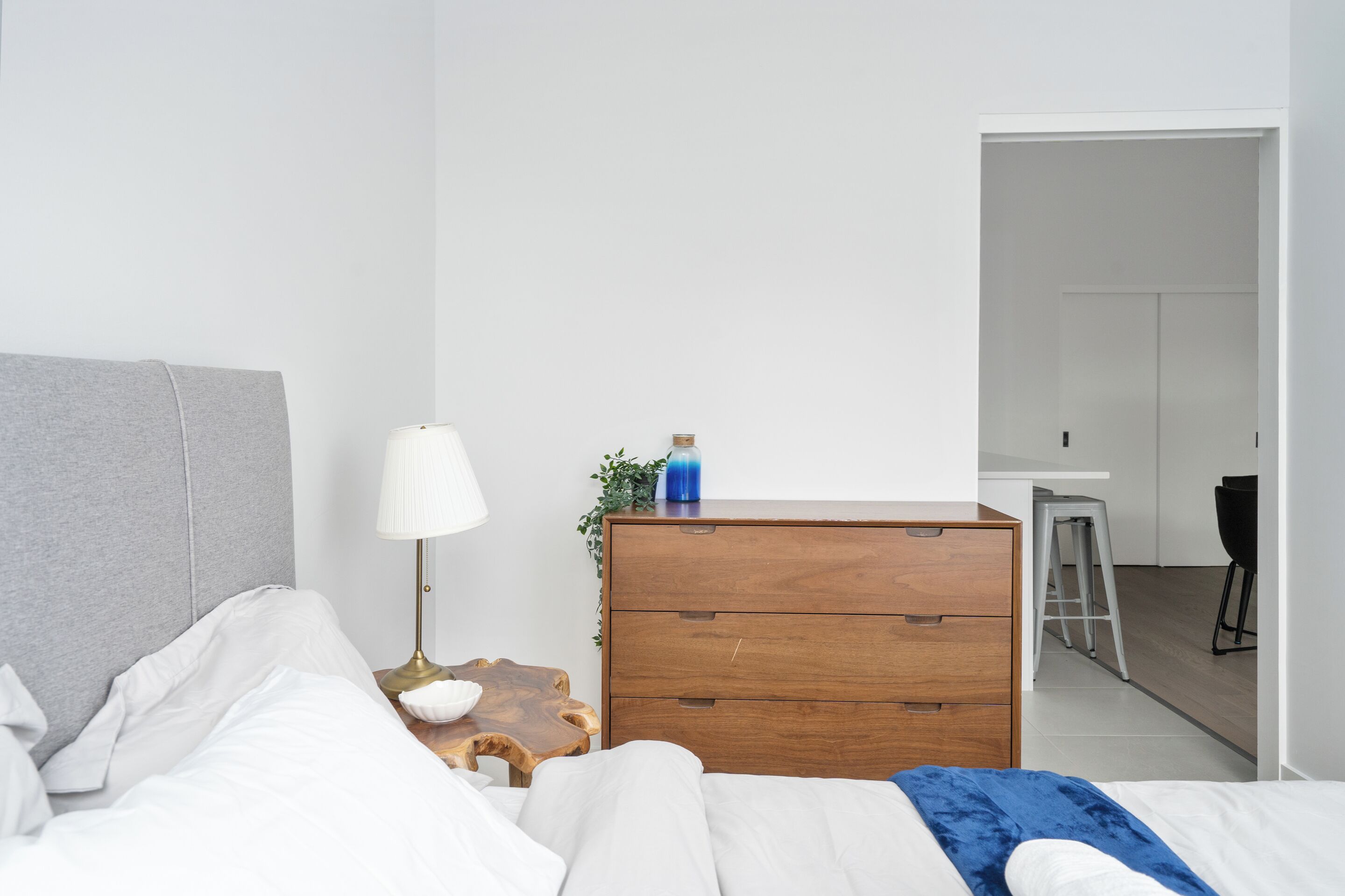 A modern bedroom with a bed, bedside table, lamp, dresser, and a view into the adjacent bathroom.