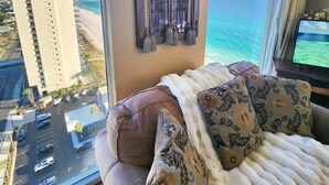 Ein Zimmer mit einem großen Fenster, das Blick auf den Strand und ein hohes Gebäude gewährt. Es gibt ein Sofa mit gemusterten Kissen und einer weißen Tagesdecke.