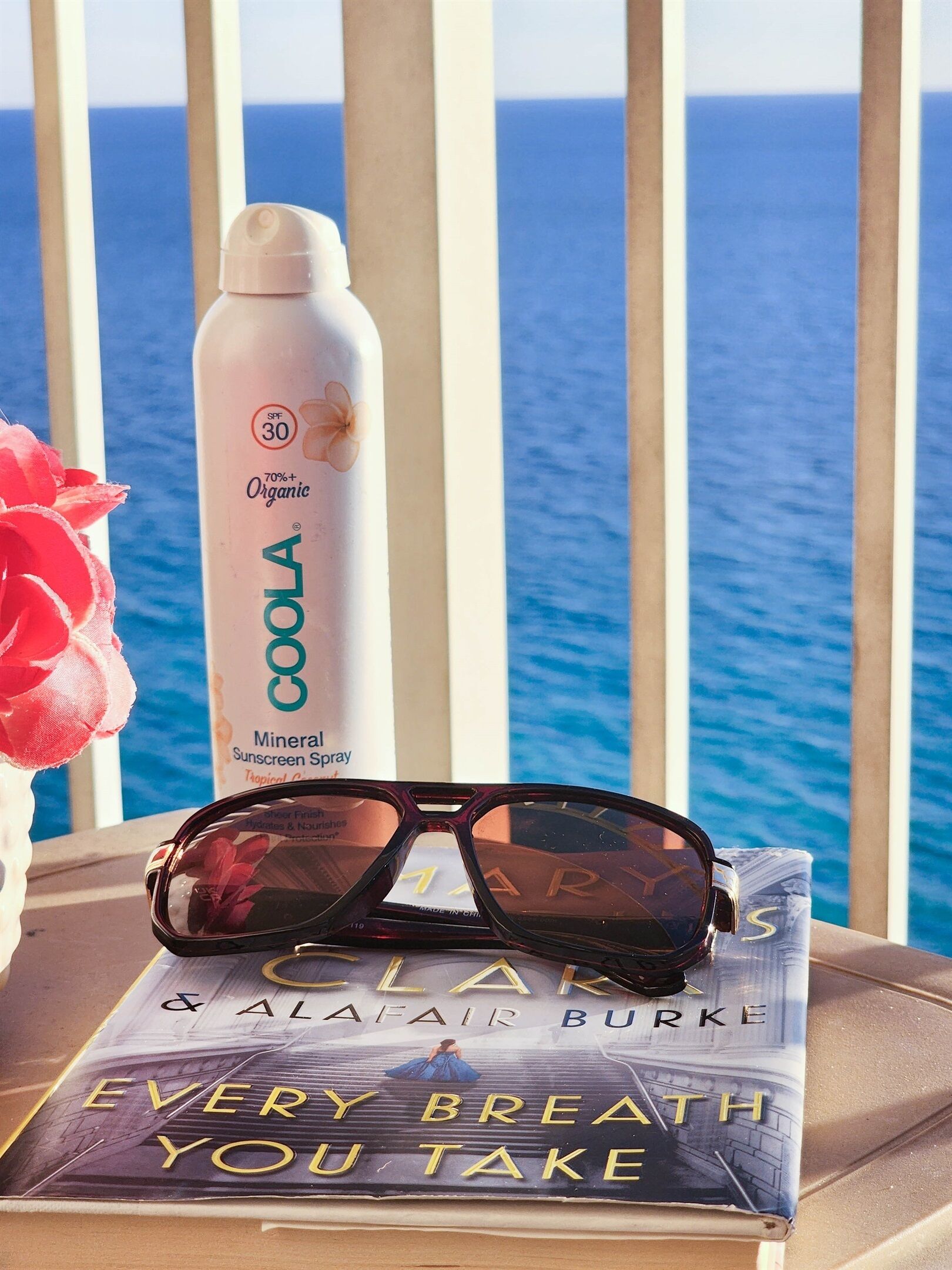 A sunscreen bottle, sunglasses, and a book titled "EVERY BREATH YOU TAKE" on a table with a view of the ocean.