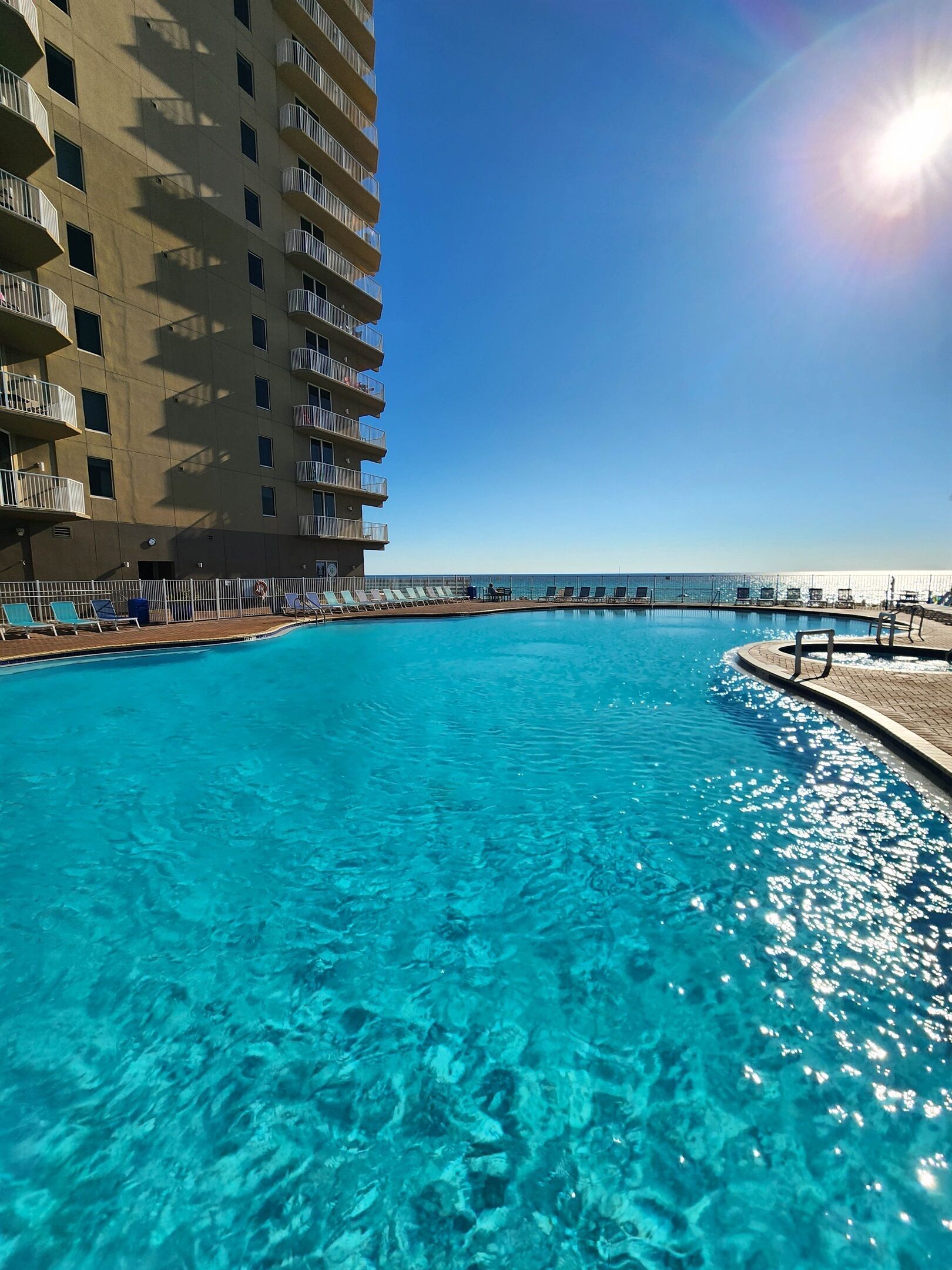 Una piscina azul cristalina con un cielo iluminado por el sol, un edificio de gran altura con balcones, y una vista distante del océano.