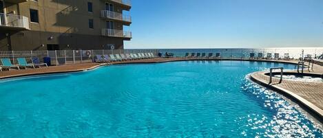 Una piscina azul cristalina con un cielo iluminado por el sol, un edificio de gran altura con balcones, y una vista distante del océano.