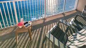 Ein Balkon mit Blick auf den Ozean und einem Sonnenuntergang über dem Wasser. Zwei Stühle und ein kleiner Tisch mit einem Buch und einer rosa Blume.