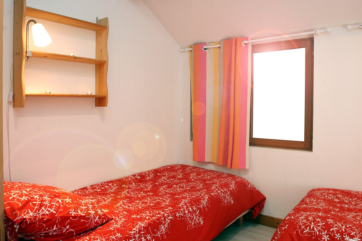 Una habitación con dos camas, un estante de madera con una lámpara, una ventana con cortinas coloridas y una pared blanca.