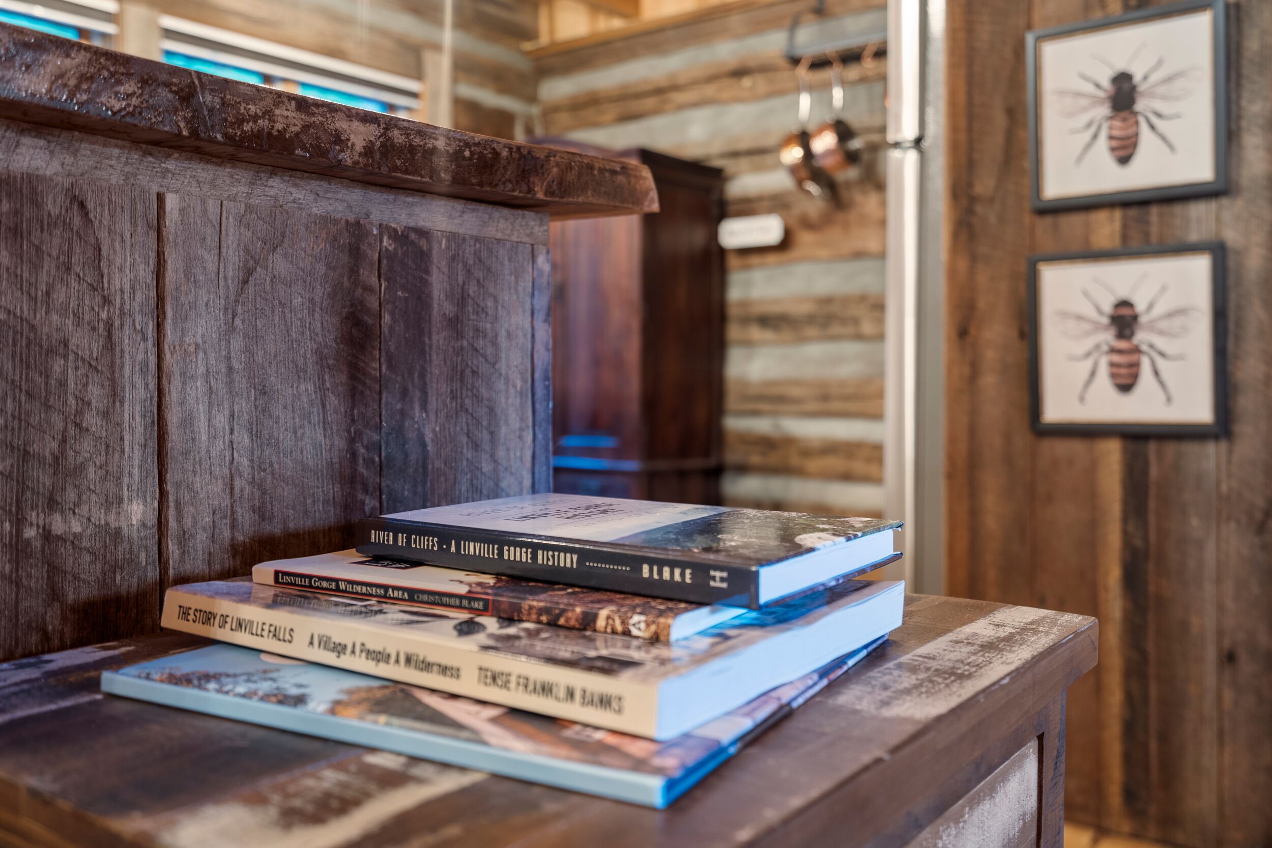 A stack of books on a wooden table in a rustic room with wooden walls and framed pictures on the wall.
