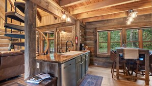 A rustic cabin interior with a wooden staircase, a kitchen with a large island, a dining area with wooden furniture, and large windows offering a view of the outdoors. - Luxury Log Cabin: Scenic Views Near Linville Falls (Marion)