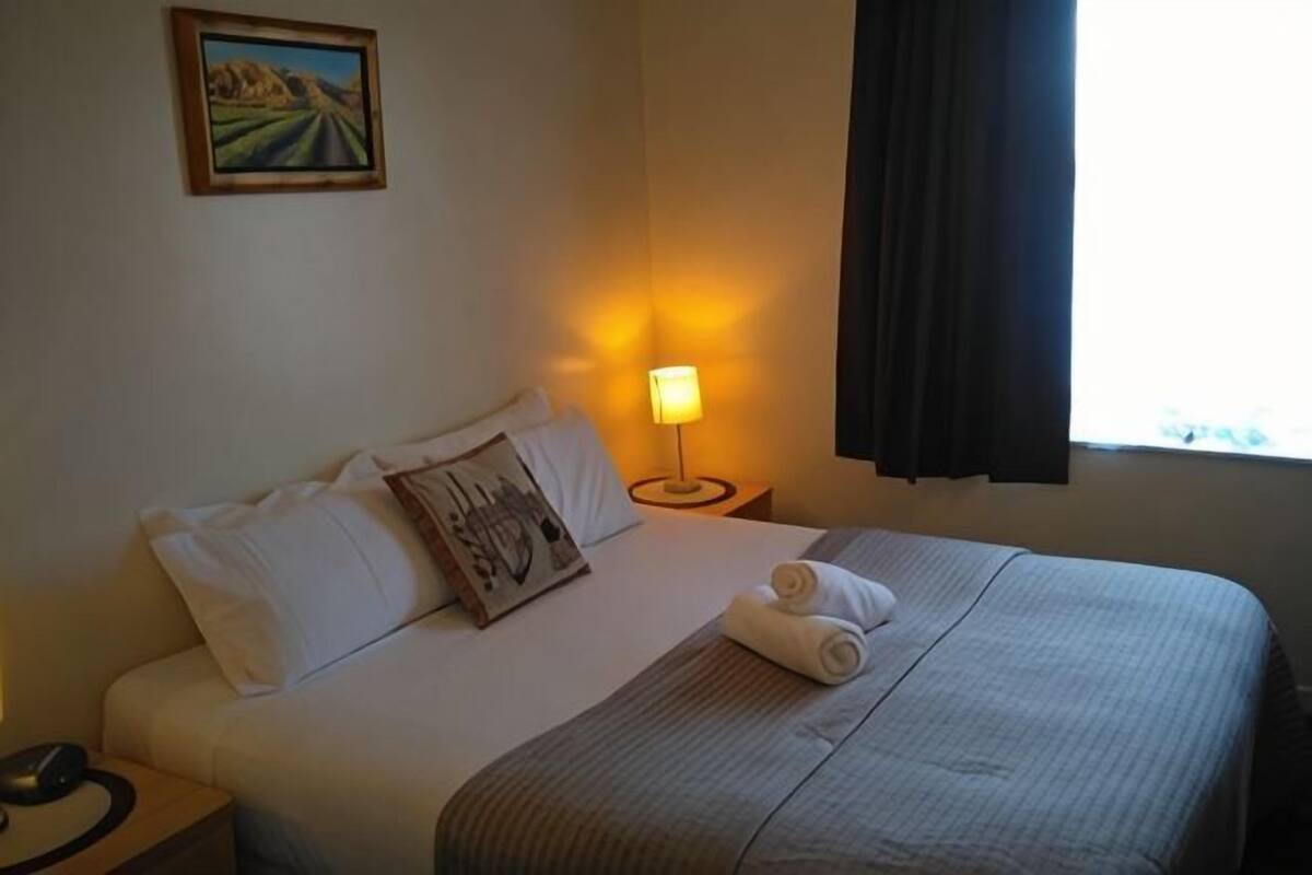 Habitación de hotel con cama, lámpara de noche, un cuadro en la pared y una ventana con cortinas.
