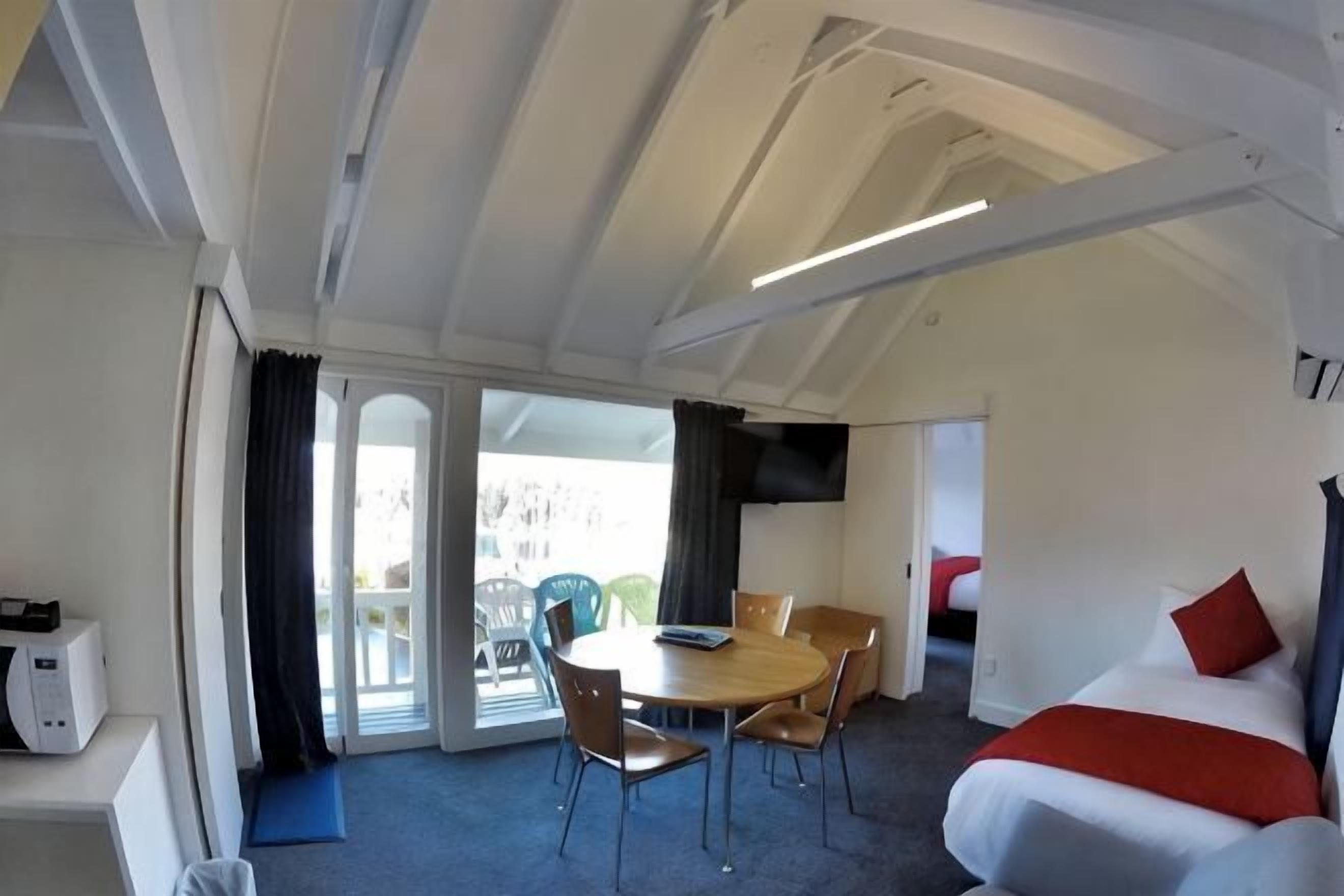 A room with a bed, a dining table with chairs, a small kitchen area, and a sliding glass door leading to an outdoor area.