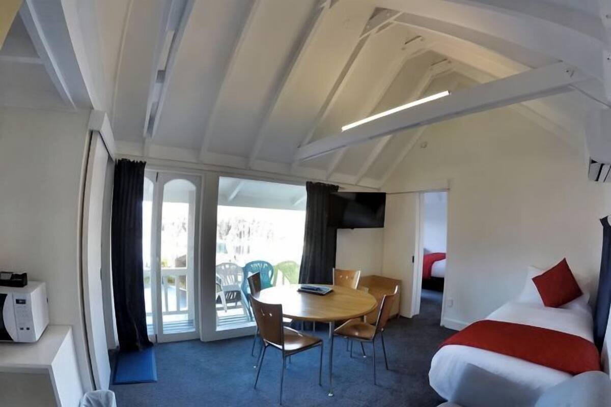Una habitación con cama, una mesa de comedor con sillas, una pequeña cocina y un ventanal corredero que da a un espacio exterior.