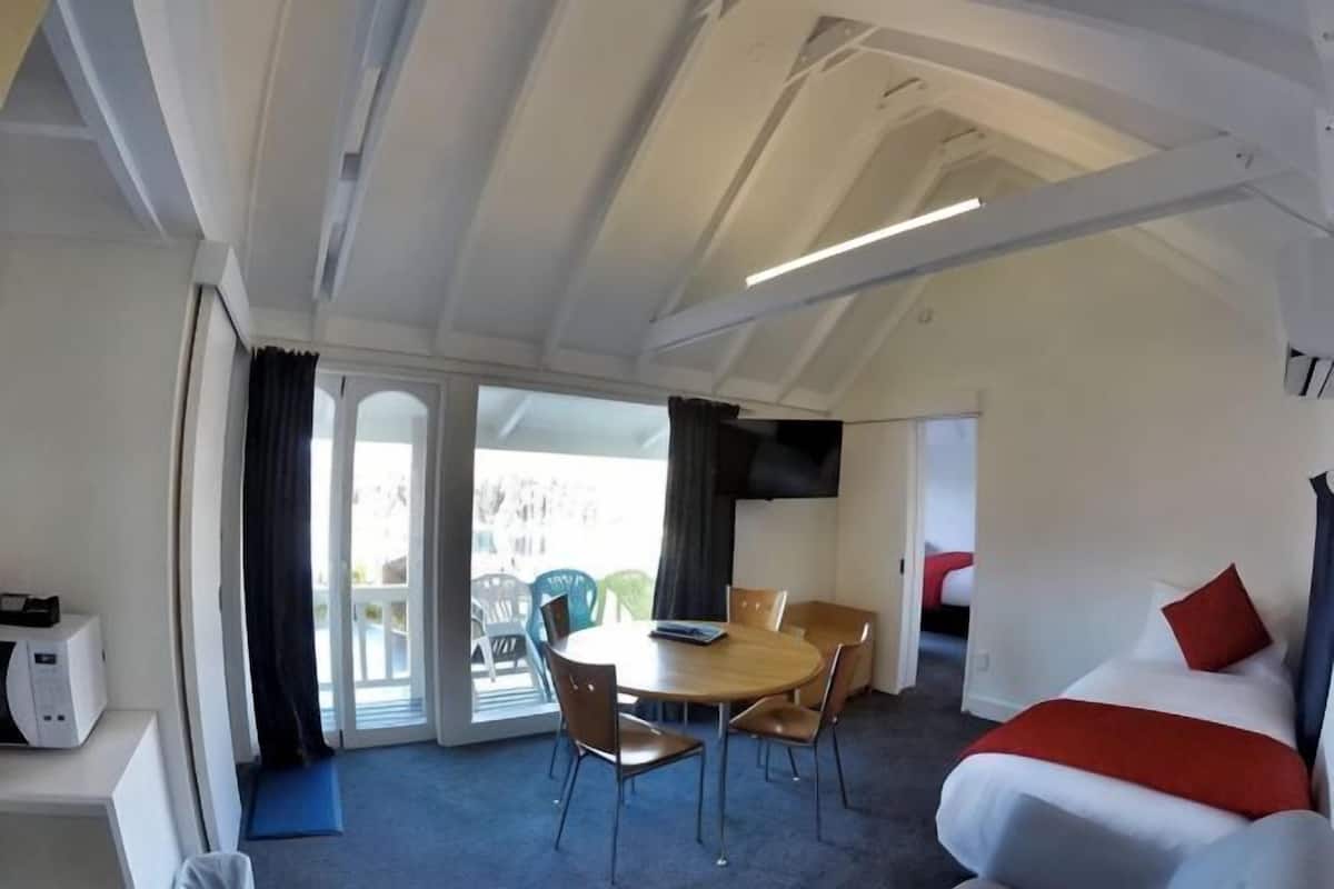 A room with a bed, a dining table with chairs, a small kitchen area, and a sliding glass door leading to an outdoor area.