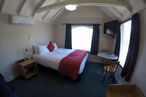 Quarto com cama, mesa de cabeceira, cadeira, mesinha, televisão e janela com cortinas.