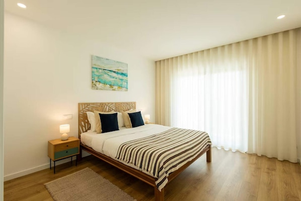 Ein Schlafzimmer mit einem hölzernen Bett, einem Nachttisch mit Lampe, einem Bild an der Wand und einem großen Fenster mit Vorhängen.