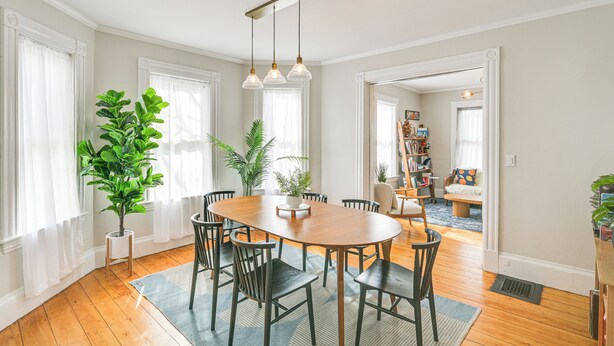Un comedor con una mesa y sillas de madera, rodeado de plantas en macetas y ventanas grandes con cortinas translúcidas.