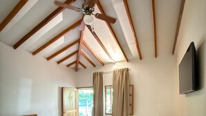 Un dormitorio con dos camas, vigas de madera, ventilador de techo, televisor y ventana con cortinas.