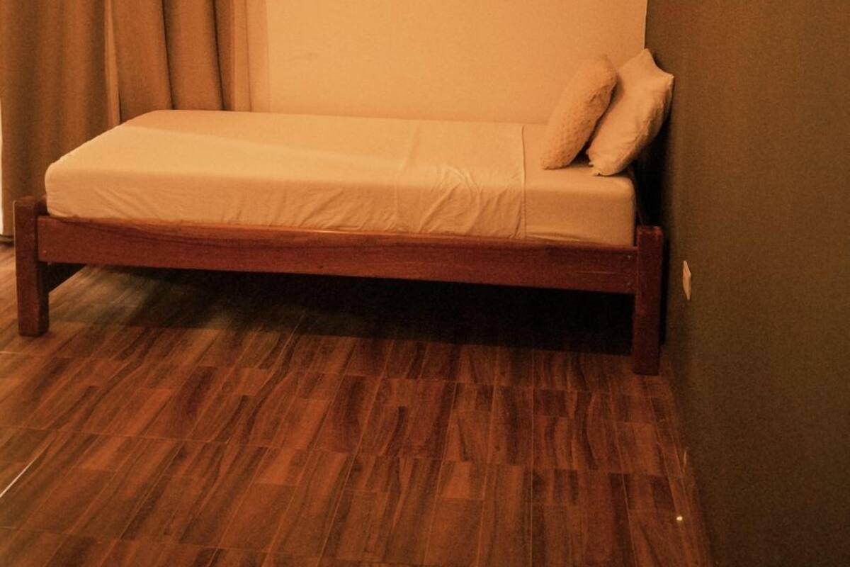 Una cama individual en una habitación con suelo de madera, pared lisa y una cortina.