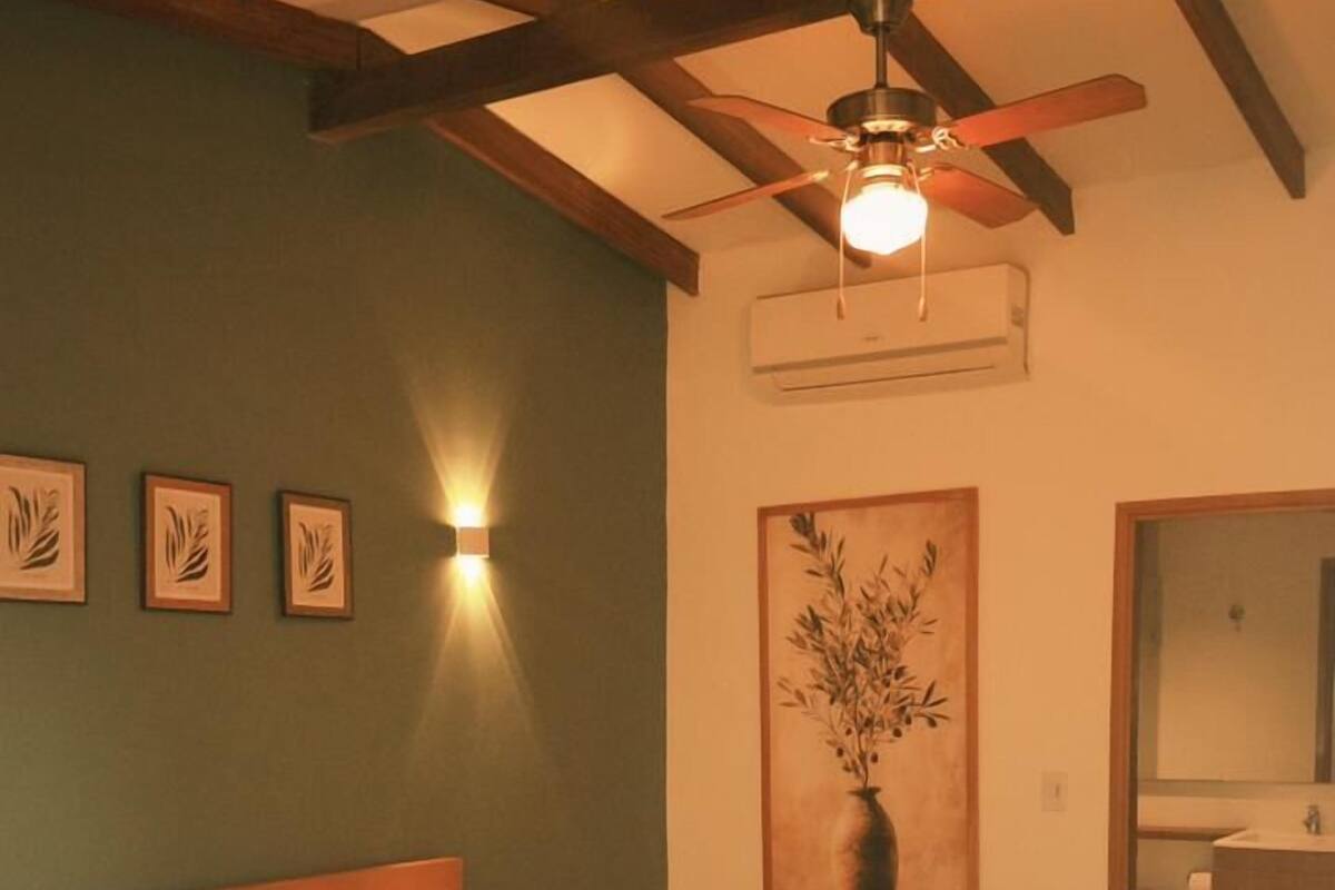 Un dormitorio con estructura de cama de madera, ventilador de techo, equipo de aire acondicionado, mesita de noche y espejo montado en la pared.