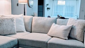 Ein Wohnzimmer mit einem grauen Ecksofa, einem Couchtisch und einer flauschigen, braunen Decke. Drei Pendelleuchten, ein Spiegel und eine Tür zu einem anderen Raum sind ebenfalls zu sehen.