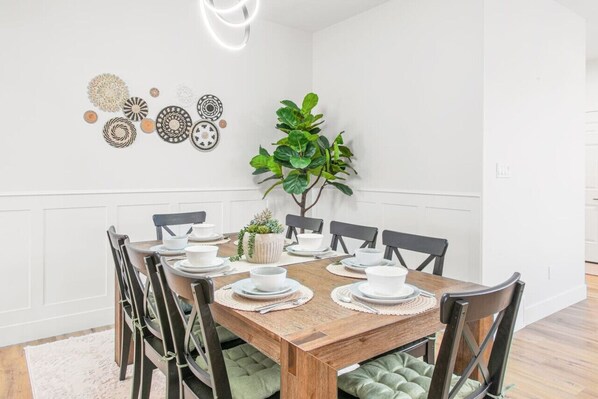 Un comedor con una mesa de madera dispuesta para seis personas, rodeada de sillas con cojines verdes. Hay una planta en maceta sobre la mesa, y platos decorativos en la pared.