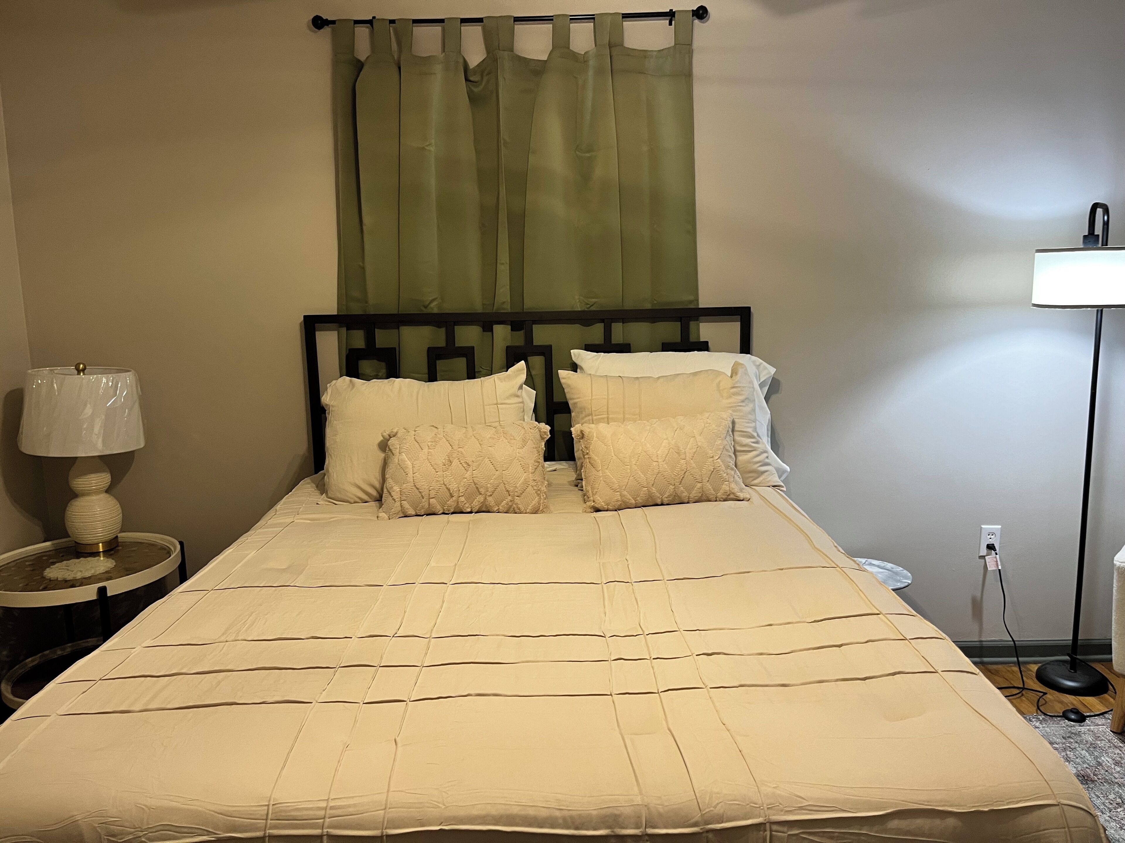 Ein ordentlich bezogenes Bett mit einer beigen Tagesdecke und Kissen, einer Nachttischlampe und einem grünen Vorhang.