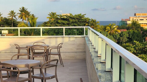 Eine Terrasse mit Tisch und Stühlen, mit Blick auf eine Küstenlandschaft mit Palmen und einem Gebäude im Hintergrund.