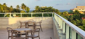 Une terrasse avec une table et des chaises, donnant sur une zone côtière bordée de palmiers et sur laquelle on aperçoit un bâtiment au loin.