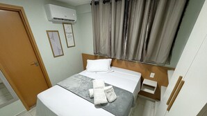 Ein ordentlich bezogenes Bett mit weißer Decke und grauer Decke, ein weißes Kopfkissen und ein Handtuch mit der Aufschrift "TEKATE". Ein Holz-Kopfseitenverkleidung, ein kleiner Nachttisch und eine Wand-Klimaanlage sind ebenfalls vorhanden.