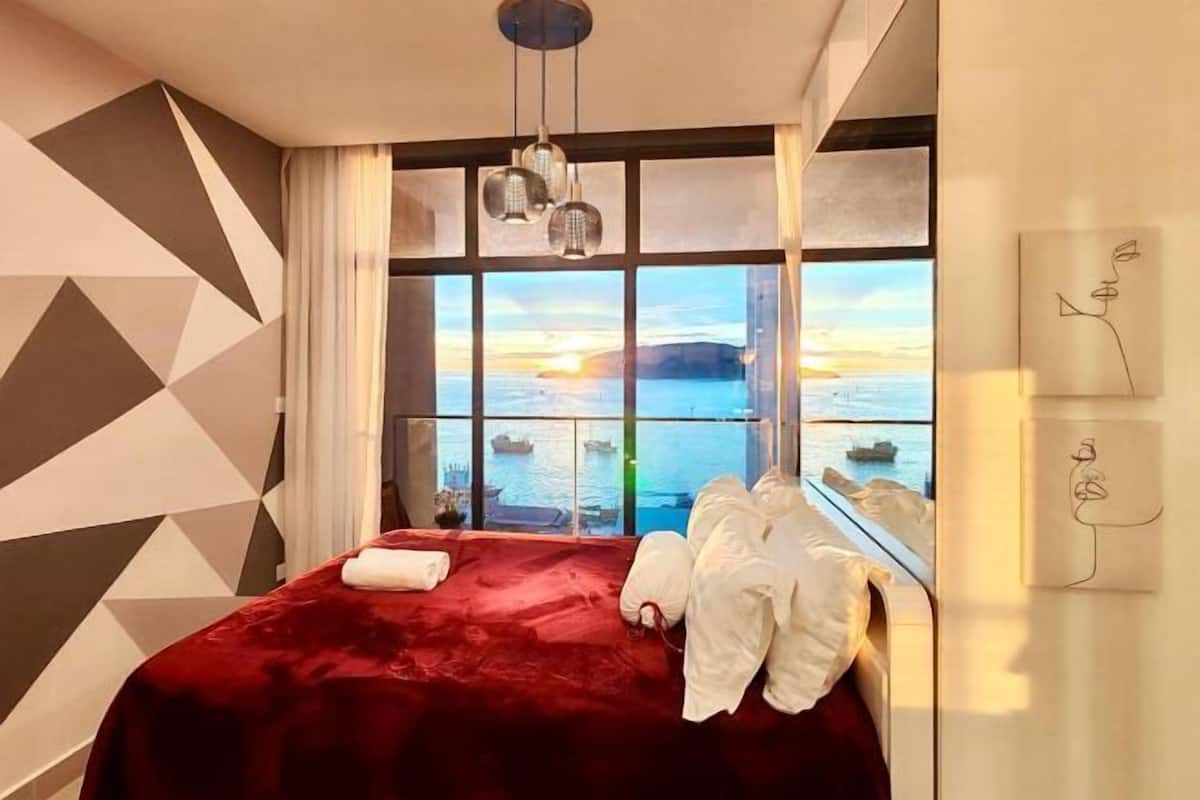 Hotelkamer met een groot raam met uitzicht op zee en boten, een rode sprei en een geometrisch wandontwerp.