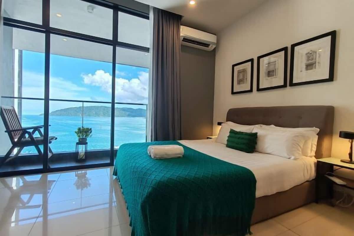 Een hotelkamer met een groot raam, een bed met een groene deken, een stoel en een nachtkastje.