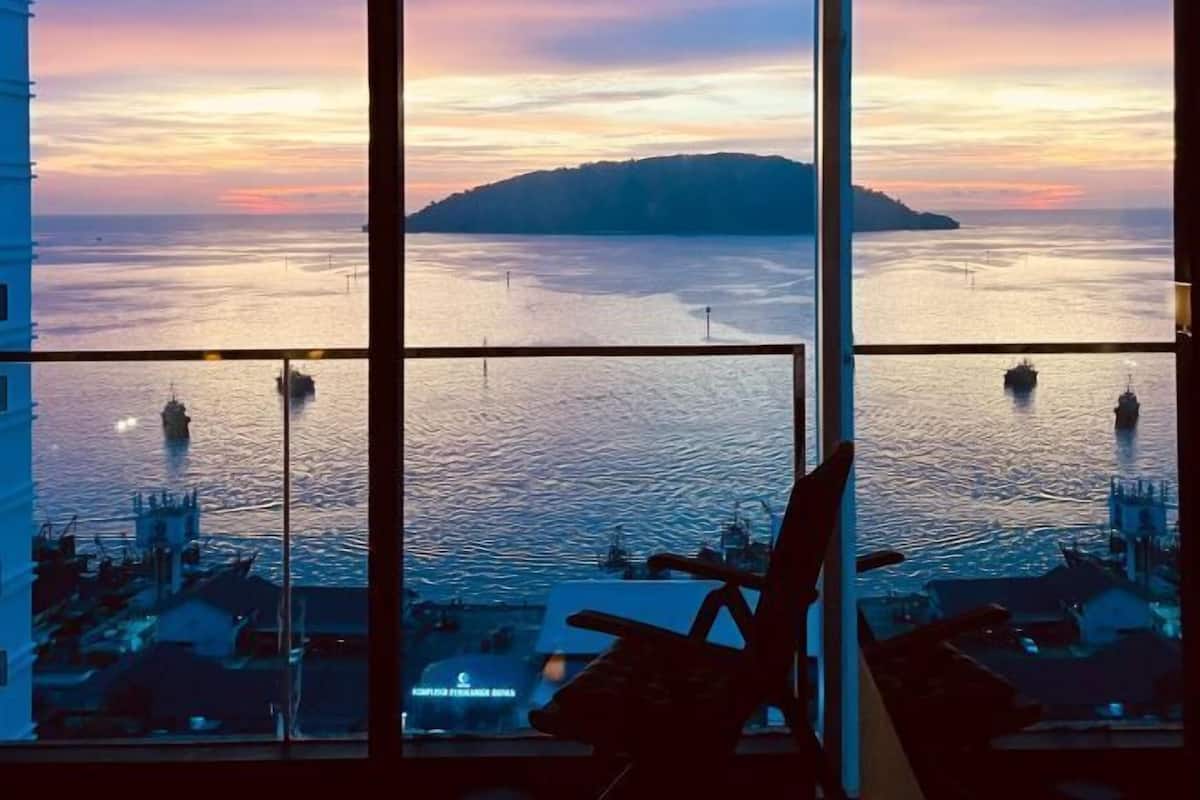 Uitzicht vanuit een hotelkamer op een hoge verdieping, over een wateroppervlak met een eiland in de verte, met een stoel en een tafel op de voorgrond.