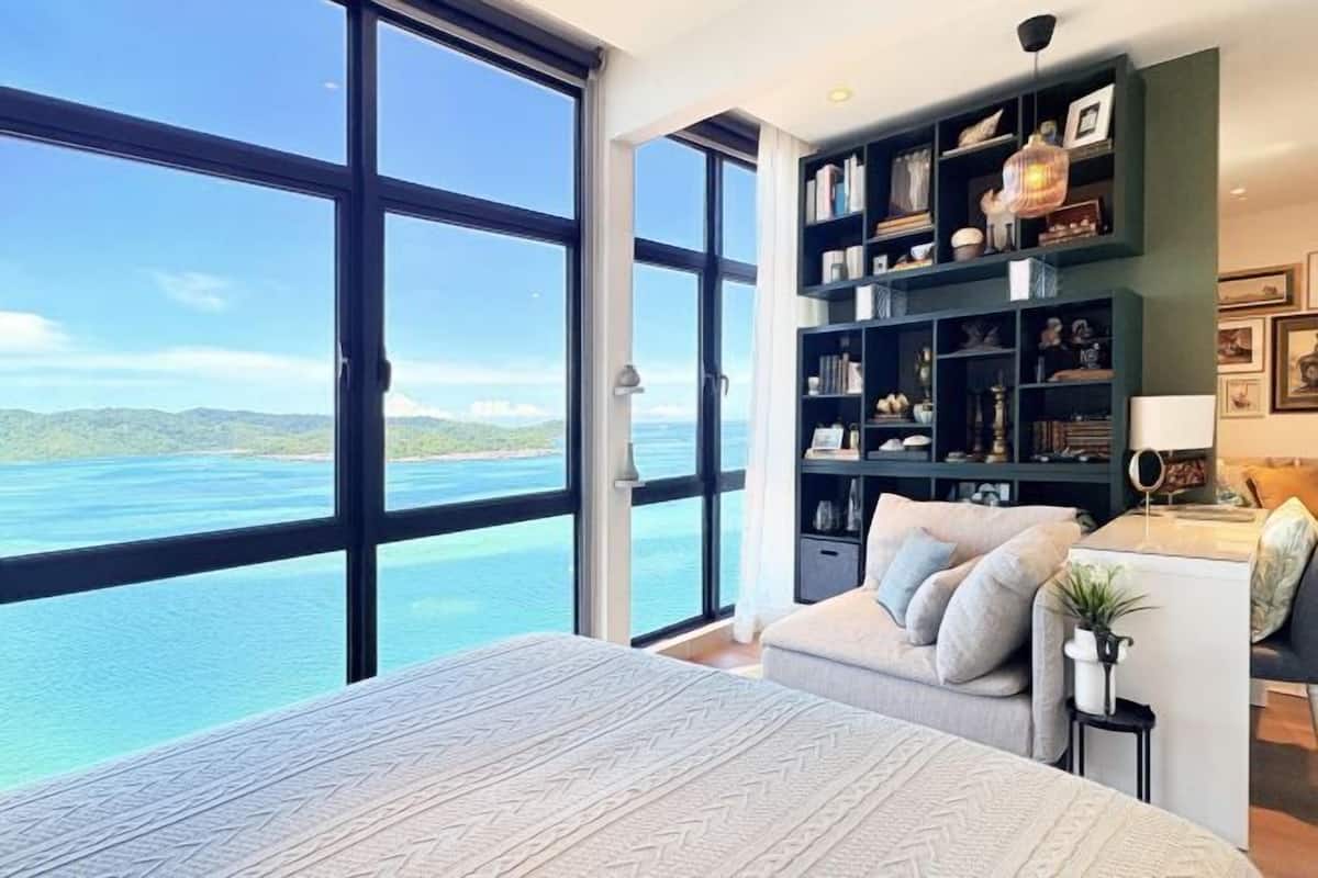 Een slaapkamer met een groot raam, een bed, een boekenkast en uitzicht op zee.