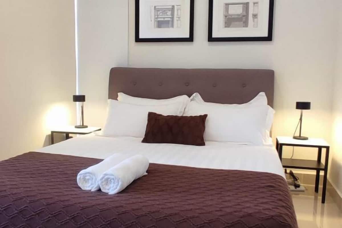 Een net opgemaakt bed met een bruine sprei en witte kussens, twee ingelijste foto’s aan de muur en nachtlampjes aan beide zijden.