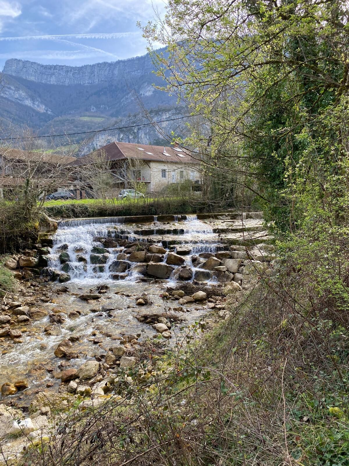 Une maison rustique avec des fondations en pierre, un petit ruisseau qui serpente à travers un lit rocheux, et un paysage montagneux en arrière-plan.