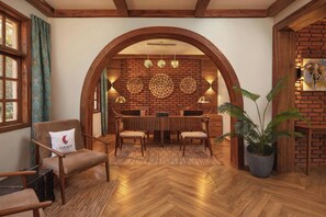 A cozy room with a wooden floor, a dining table with chairs, a sofa with a pillow, a potted plant, and a brick wall with a wooden archway. - Turaco Ngorongoro Valley, a Tribute Portfolio Lodge (Karatu)