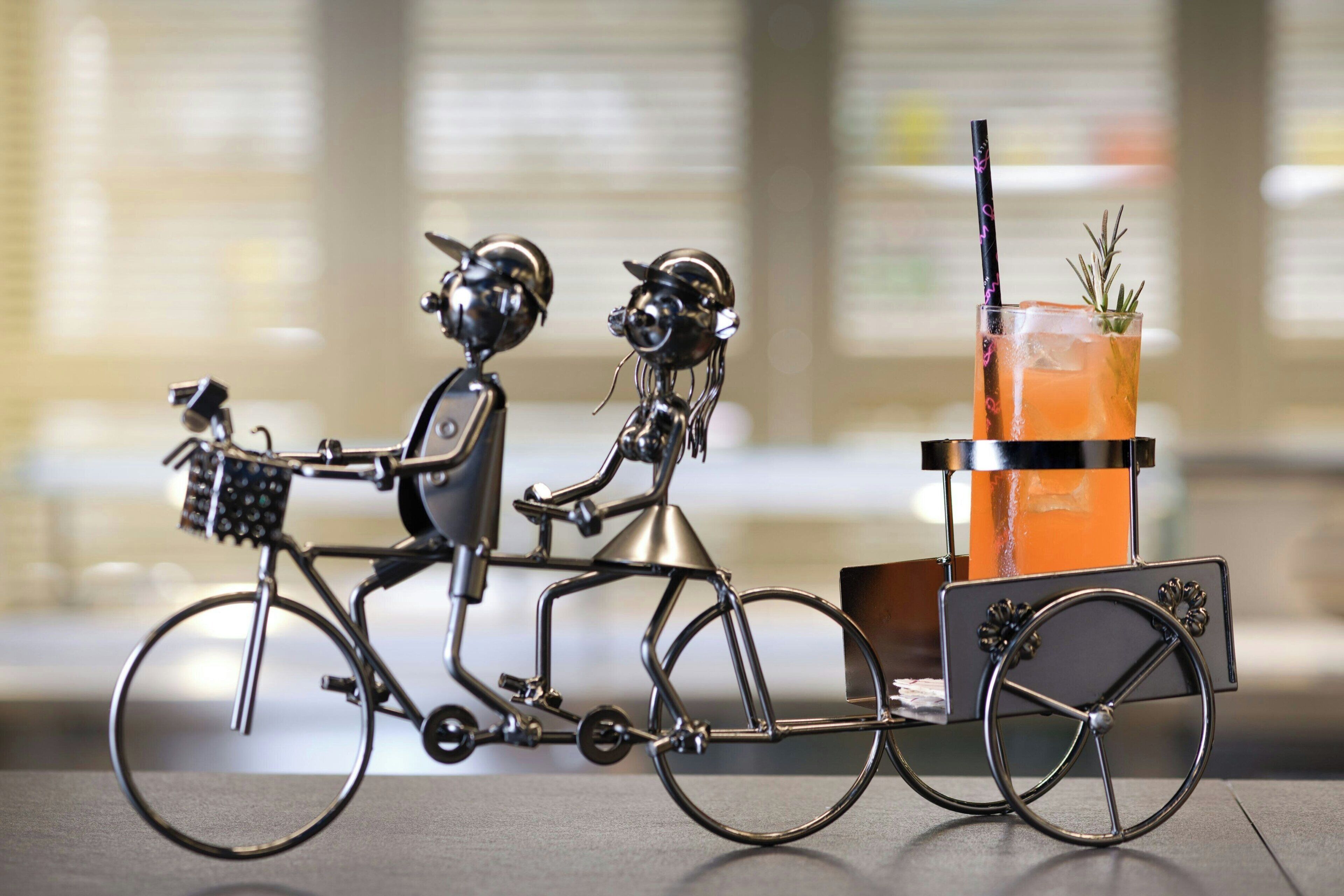Eine Metallskulptur von zwei Personen auf einem Fahrrad mit einem Wagen, der ein Getränk transportiert.