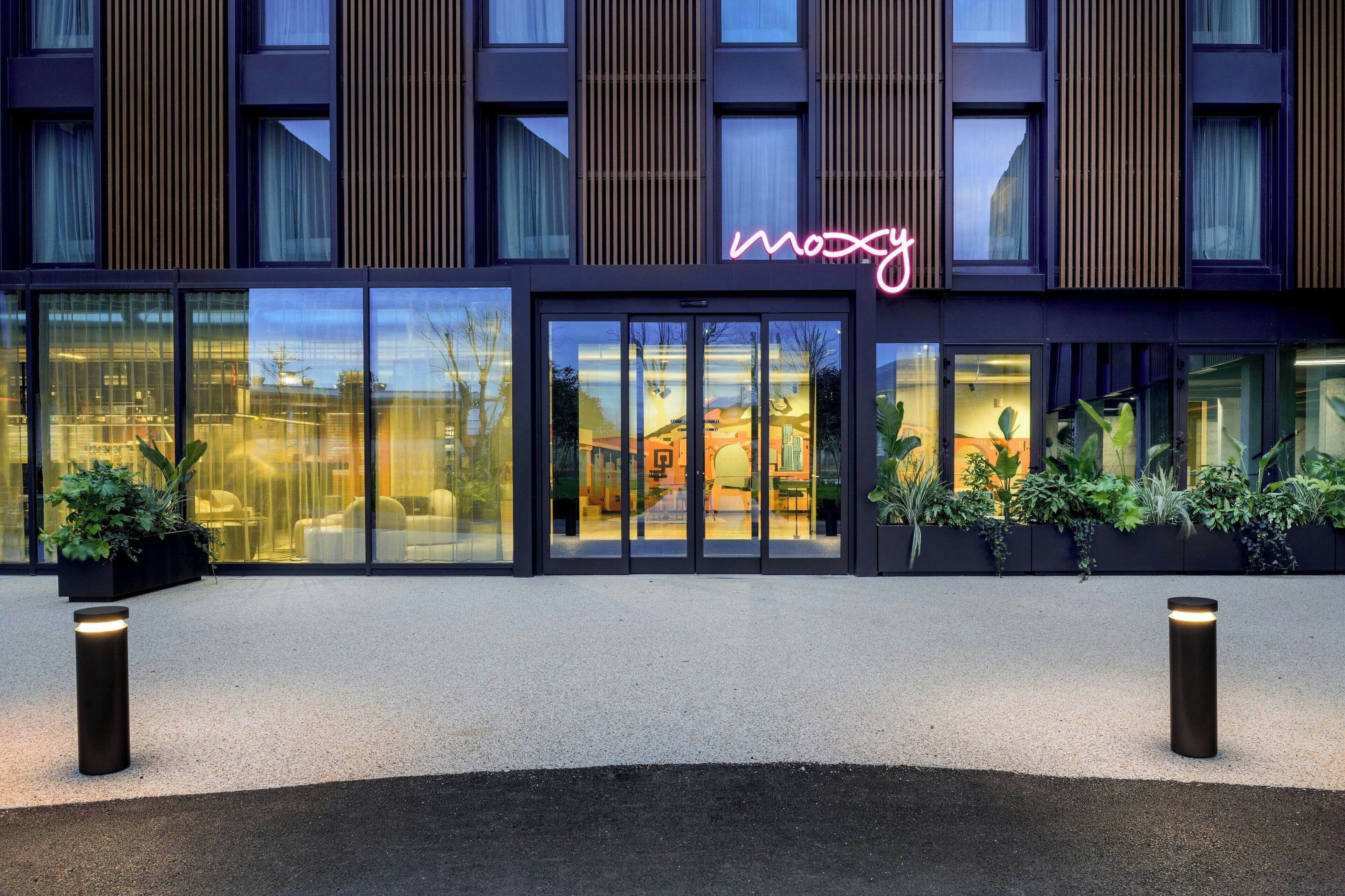 A modern hotel facade with a neon sign, large windows, and outdoor seating area.