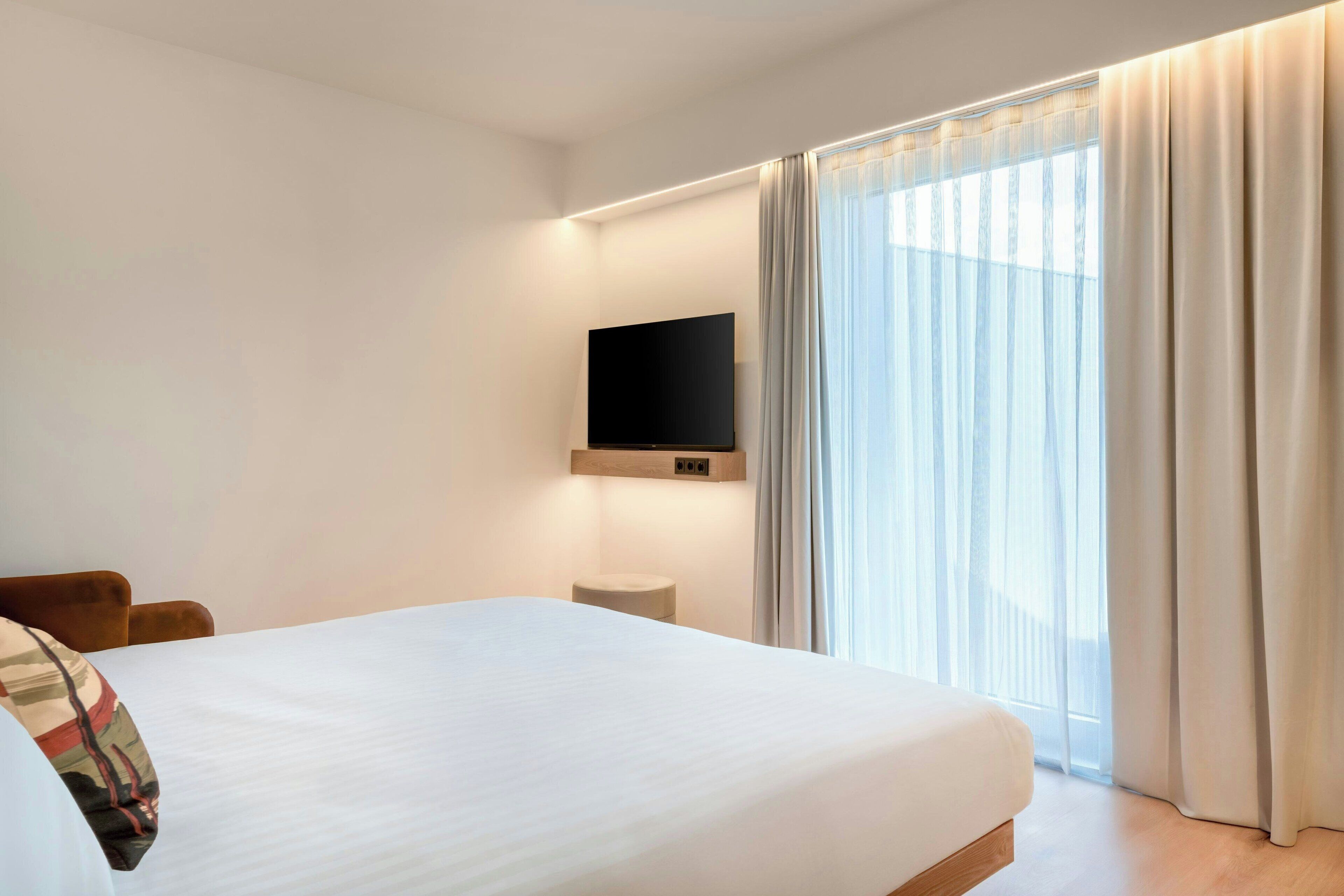 Una moderna camera d'albergo con un letto grande, una TV a schermo piatto montata a parete e un'ampia finestra con tende leggere.