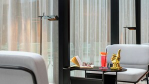 Een moderne hotelkamer met een groot raam, een klein tafeltje met een boek, glazen en een cocktail, en twee witte stoelen.