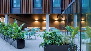 An outdoor seating area with green chairs and potted plants, a modern building with vertical wooden slats, and a glass wall reflecting the interior lights. - Moxy Pompeii (Torre Annunziata)