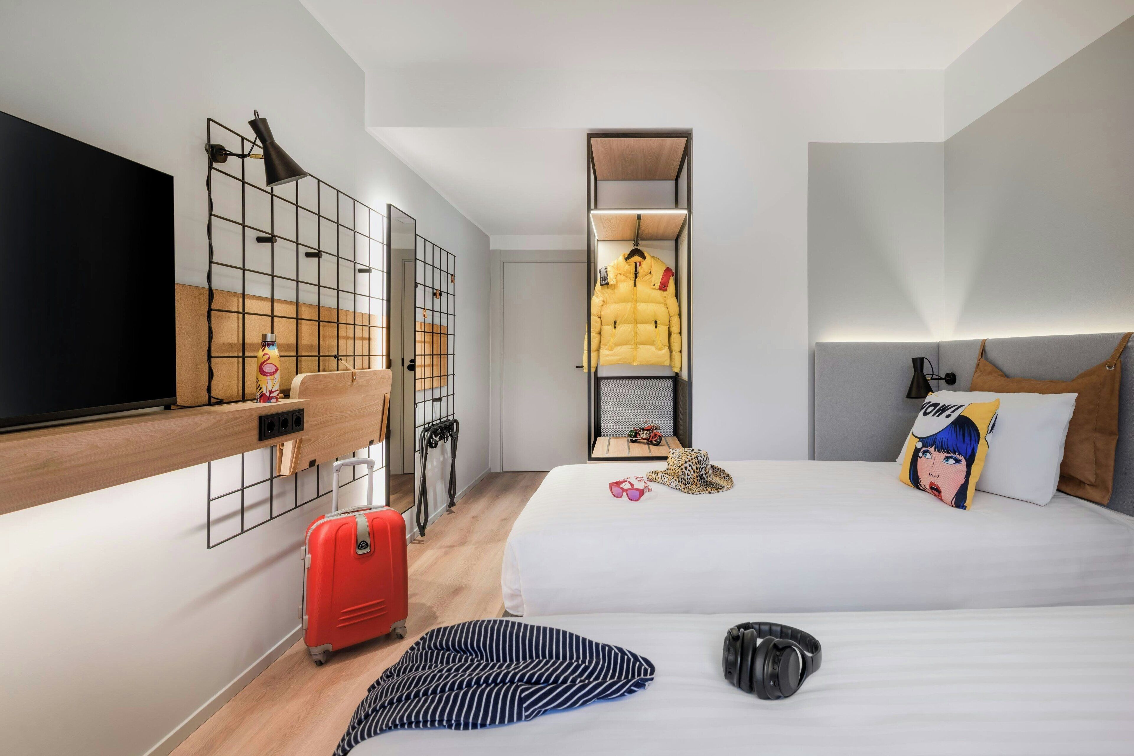 A modern hotel room with a bed, a TV, a wardrobe, a suitcase, and a small table with a bottle.