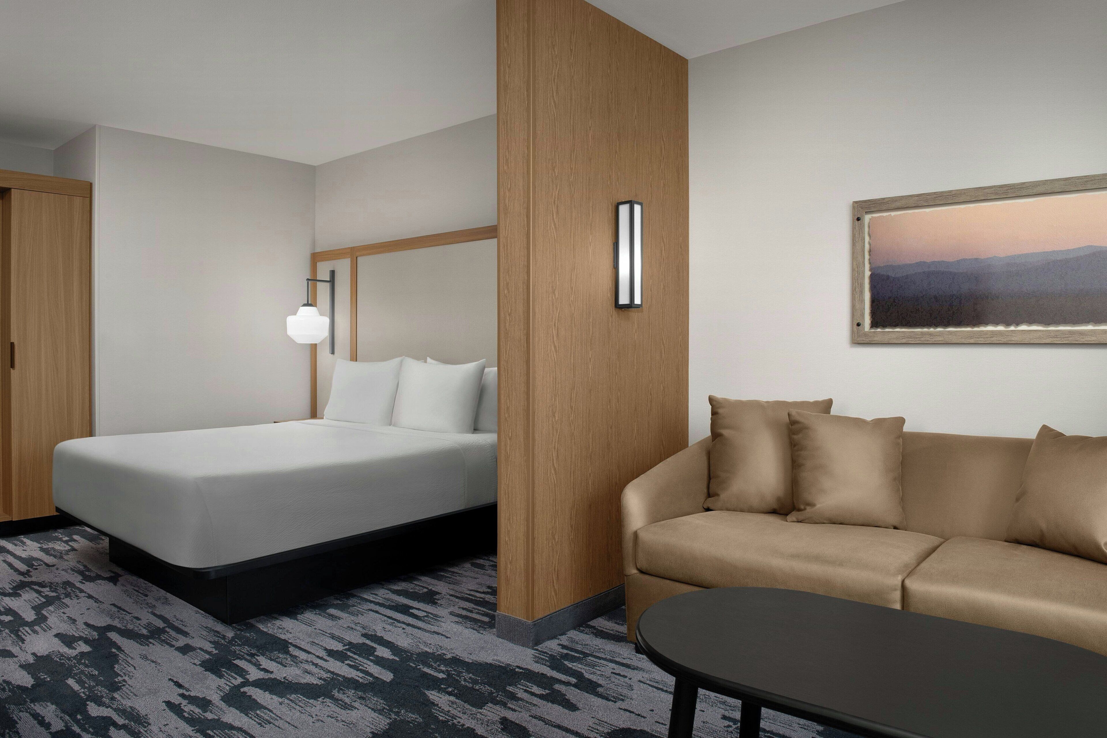 A hotel room with a bed, a sofa, a coffee table, a wall-mounted light, and a framed picture.