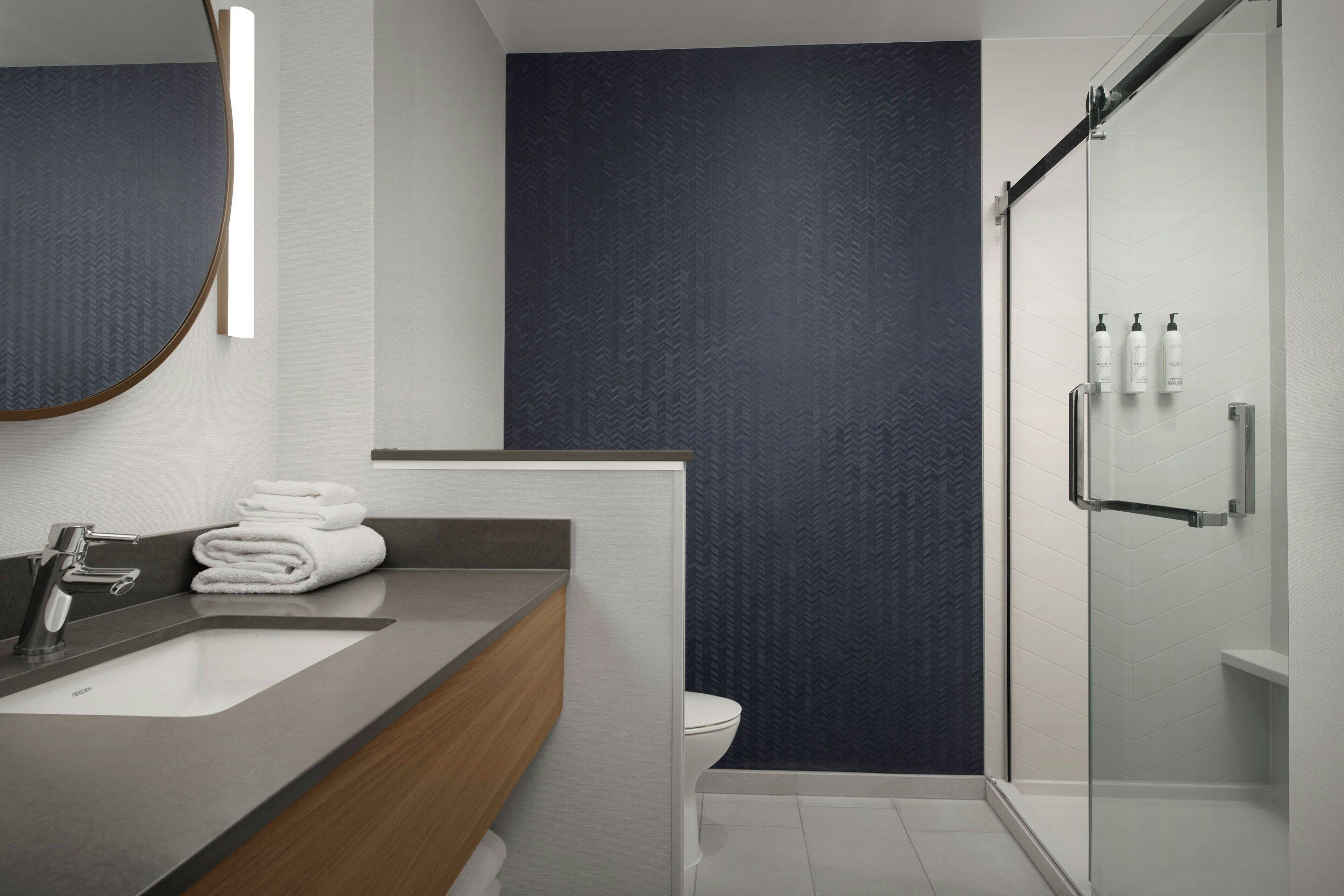 A modern bathroom with a grey countertop, a wooden vanity, a white sink, a toilet, and a glass-enclosed shower.