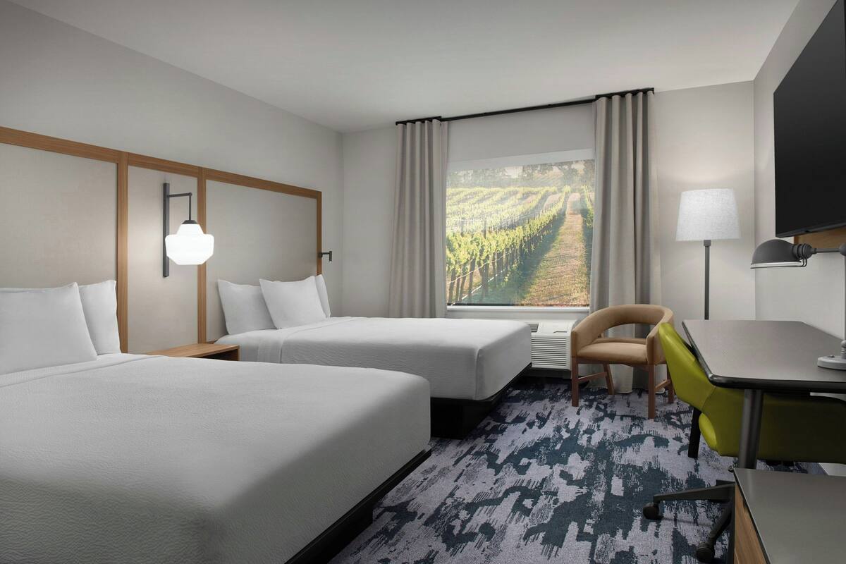 A hotel room with two beds, a desk, a chair, a TV, and a window with a vineyard view.