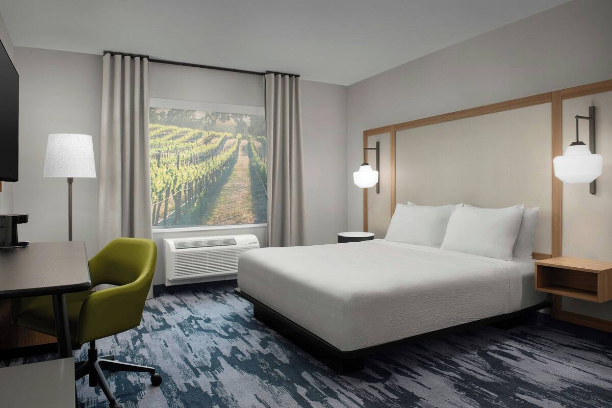 A hotel room with a bed, a desk, a chair, a window with curtains, and a view of a vineyard.