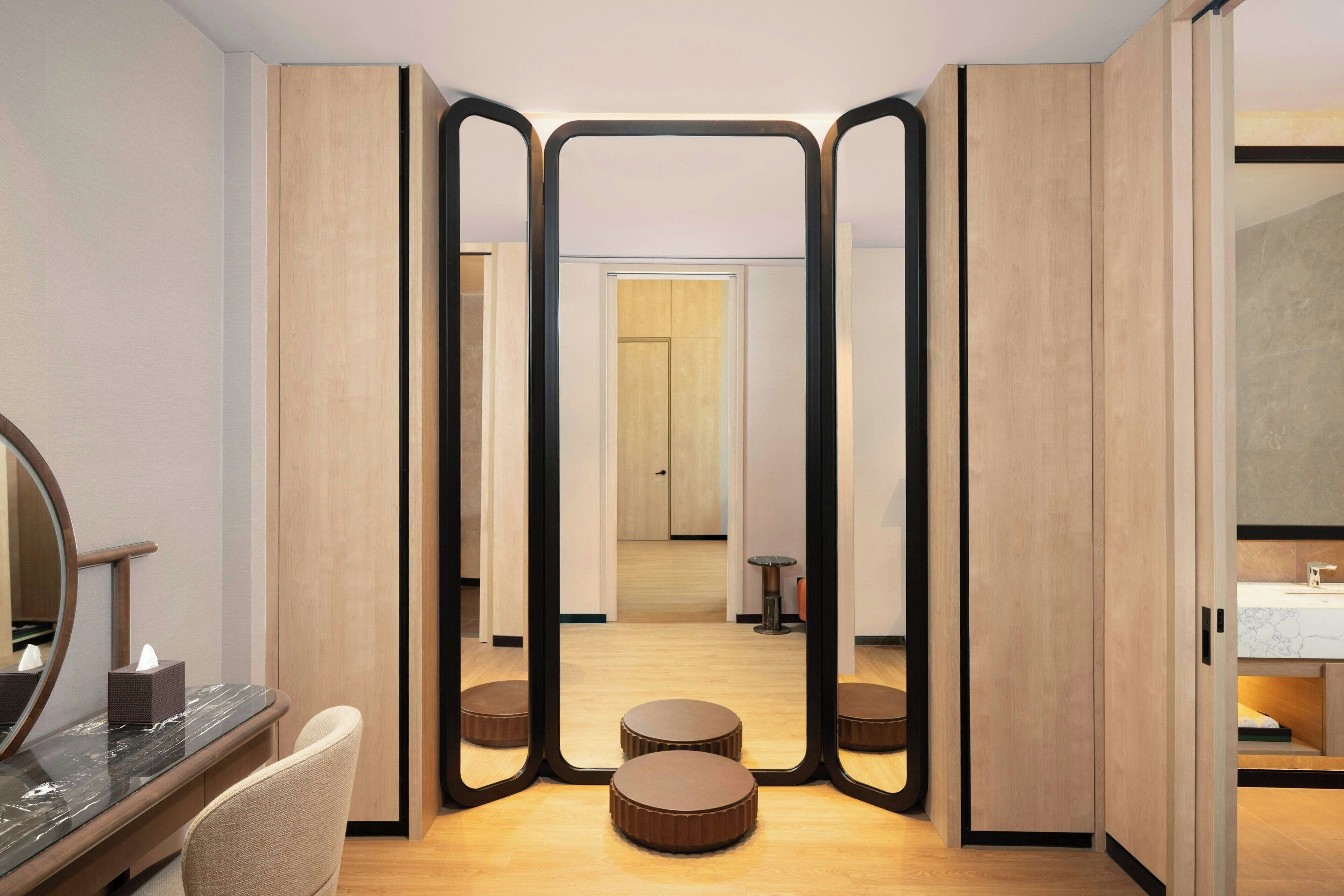Een moderne hotelkamer met een houten vloer, een ronde spiegel, een rond tafeltje en een houten kledingkast.