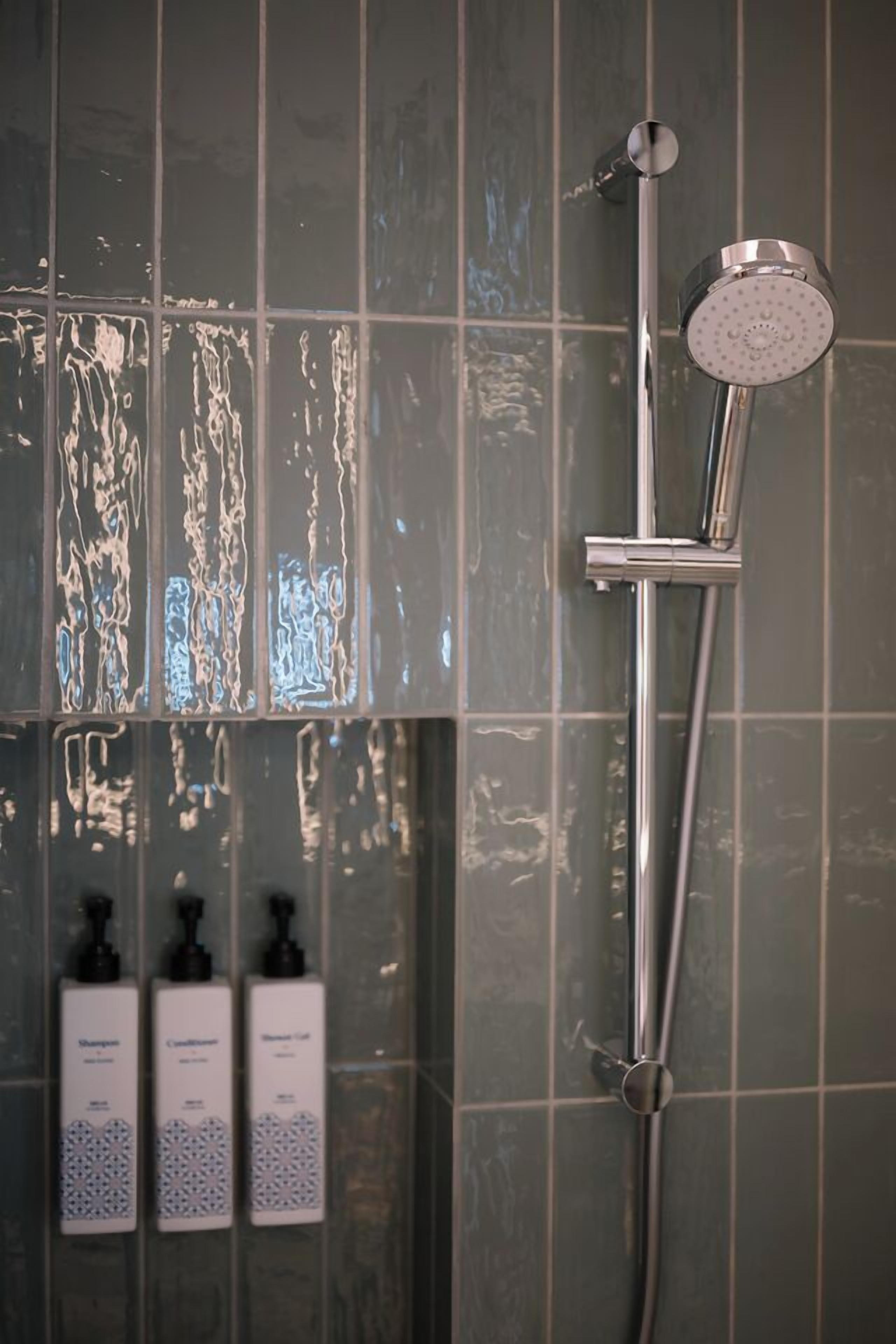 Una doccia con soffione a mano e rubinetto montato a parete, su uno sfondo di piastrelle con tre flaconi di shampoo e balsamo.