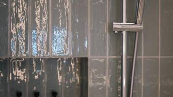 A shower with a handheld showerhead and a wall-mounted faucet, set against a tiled wall with three bottles of shampoo and conditioner.