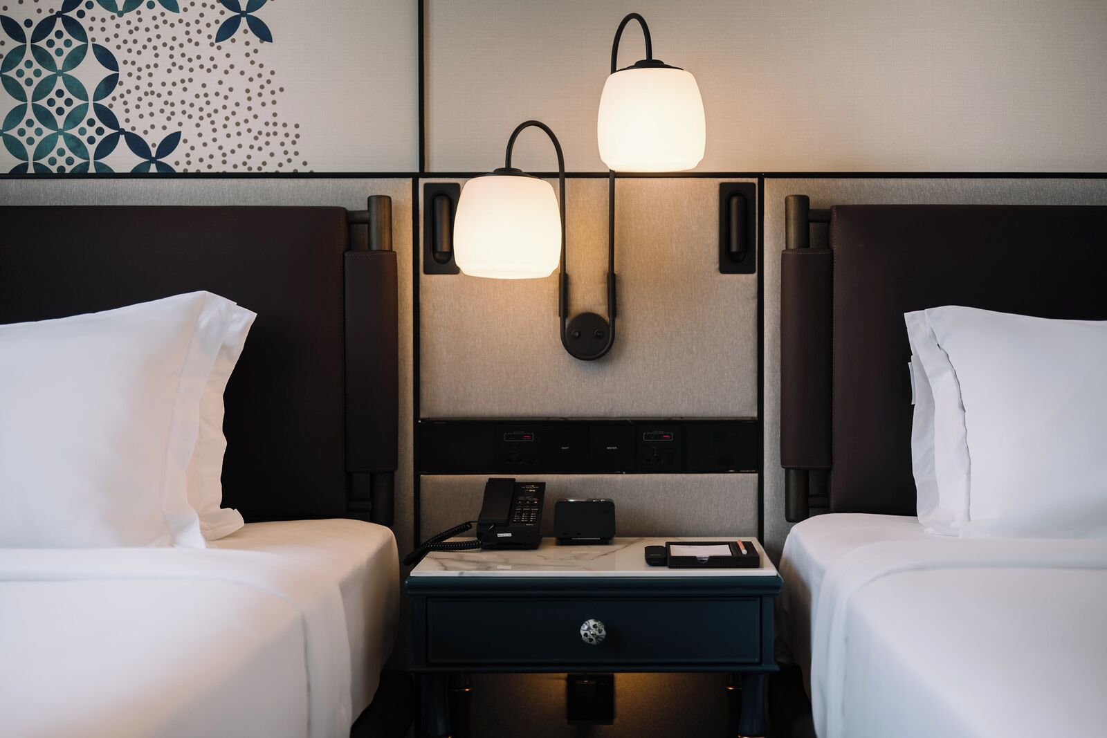 A hotel room with two beds, a nightstand with a phone and a lamp, and a wall-mounted light fixture.